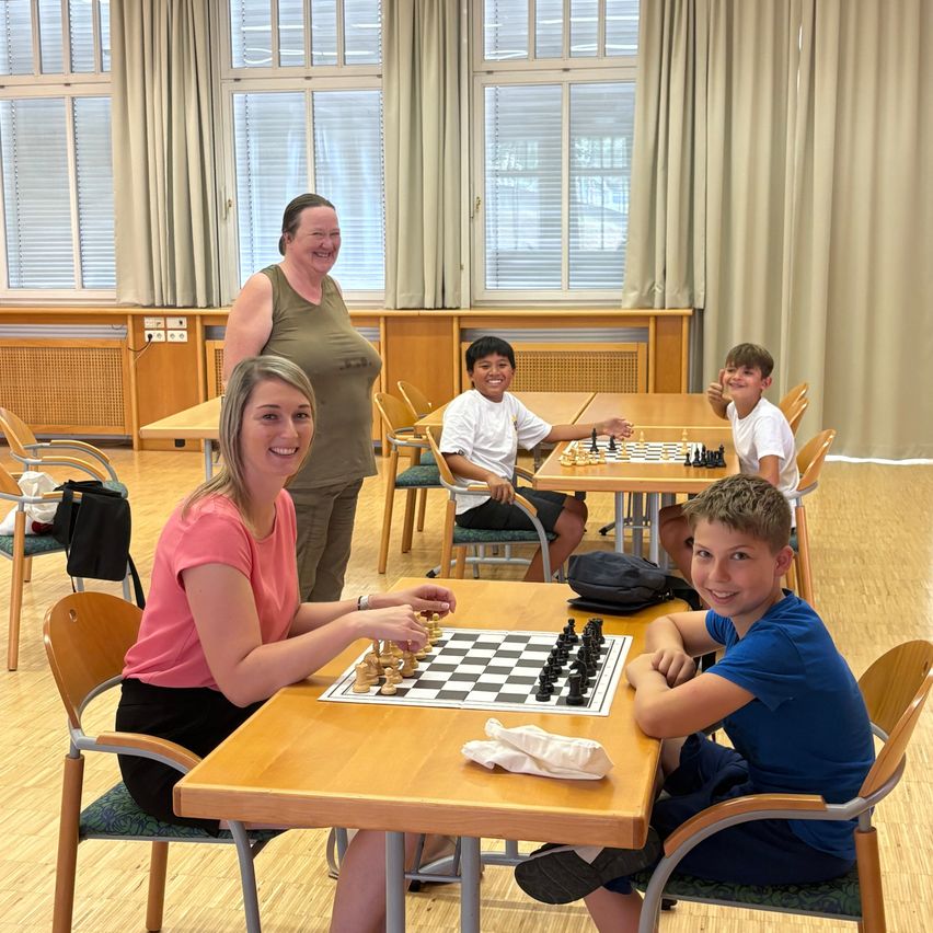 Eine Gruppe von Kindern und Erwachsenen spielt Schach in einem Raum mit Holzböden und großen Fenstern. Eine Frau in einem rosa Shirt sitzt an einem Tisch mit einem Jungen in einem blauen Shirt. Hinter ihnen spielen zwei Jungen Schach an einem anderen Tisch.