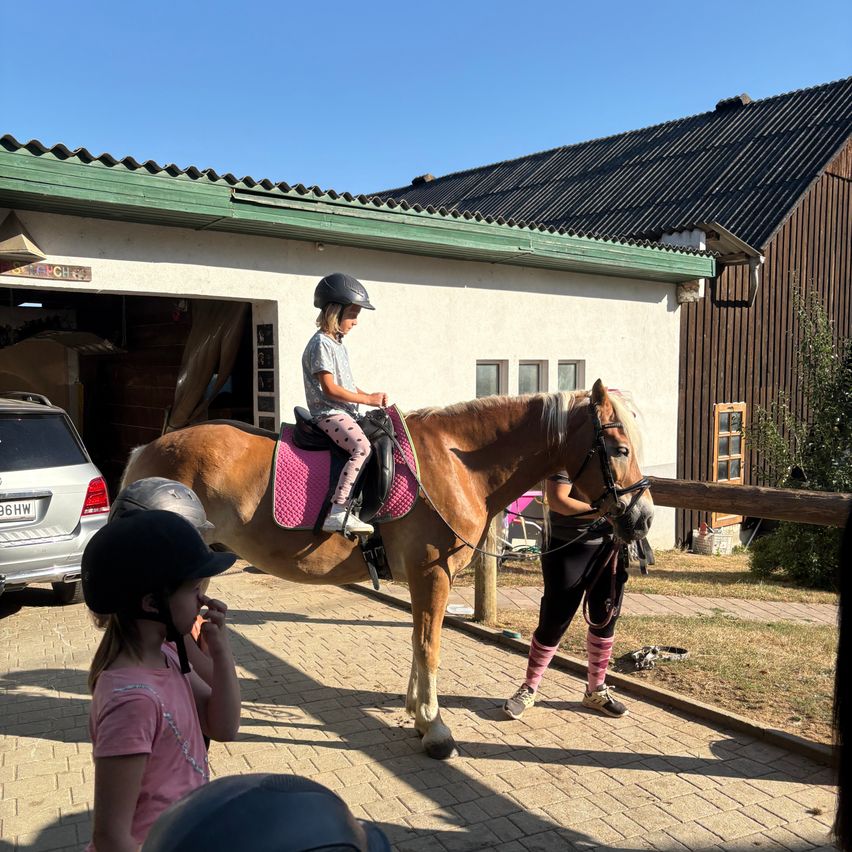 A young girl rides a brown horse with a pink saddle in front of a building. Another girl in pink socks watches from the side.