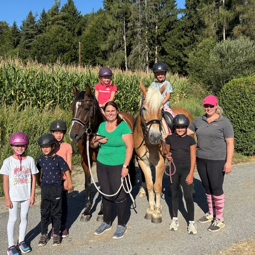 A group of people, including children, stand with two horses in a rural setting. The horses are led by two women, one holding the lead ropes. The children are wearing helmets and riding the horses. The background features a cornfield and trees.