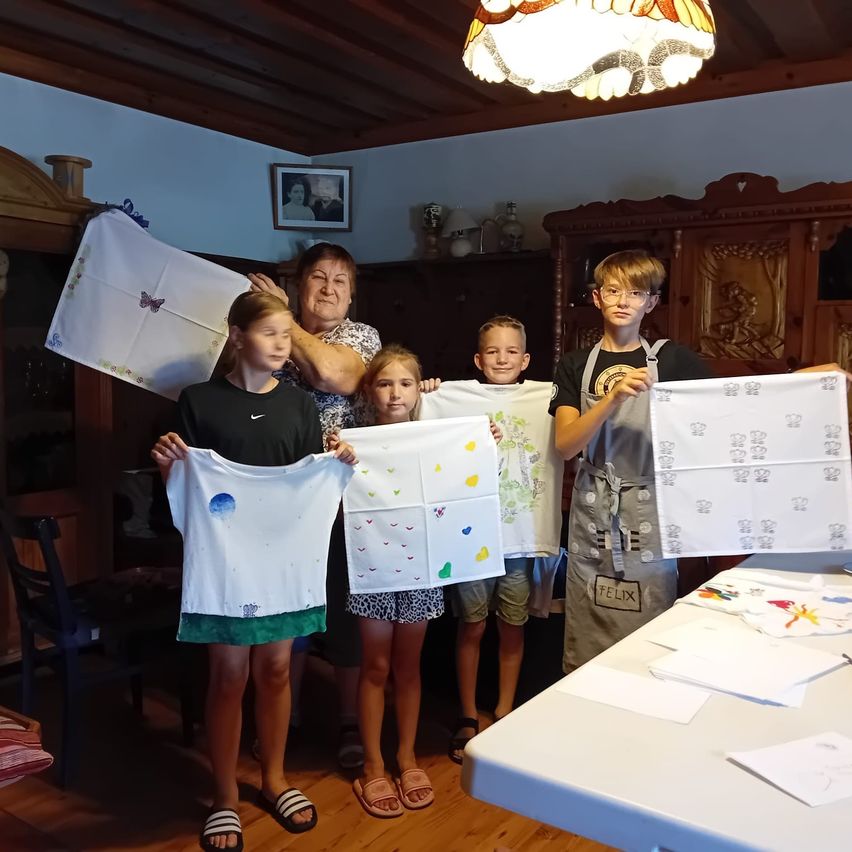 Eine Gruppe von Kindern und eine ältere Frau stehen in einem Raum und halten weiße Tücher mit Zeichnungen hoch. Der Raum hat Holzböden und Möbel, und eine Lampe hängt von der Decke.