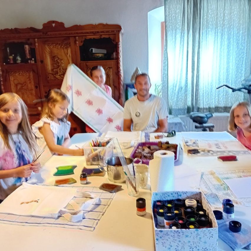 Eine Gruppe von Kindern und einem Erwachsenen versammelt sich um einen Tisch und ist in ein Kunstprojekt vertieft. Auf dem Tisch sind verschiedene Kunstmaterialien wie Farbe, Pinsel und Papier verteilt. Die Kinder lächeln und scheinen Spaß zu haben.