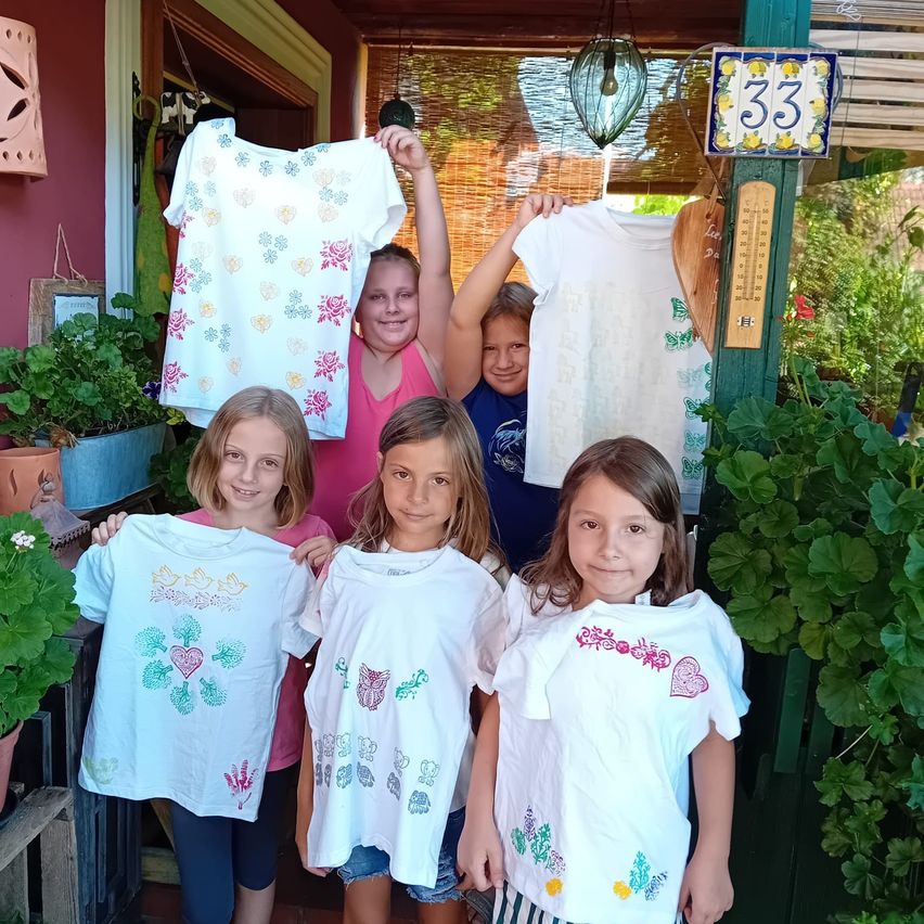 Fünf junge Mädchen lächeln und posieren für ein Foto, während sie ihre weißen T-Shirts mit verschiedenen Designs hochhalten. Sie stehen vor einem Haus mit Pflanzen und einem Schild mit der Nummer 33.