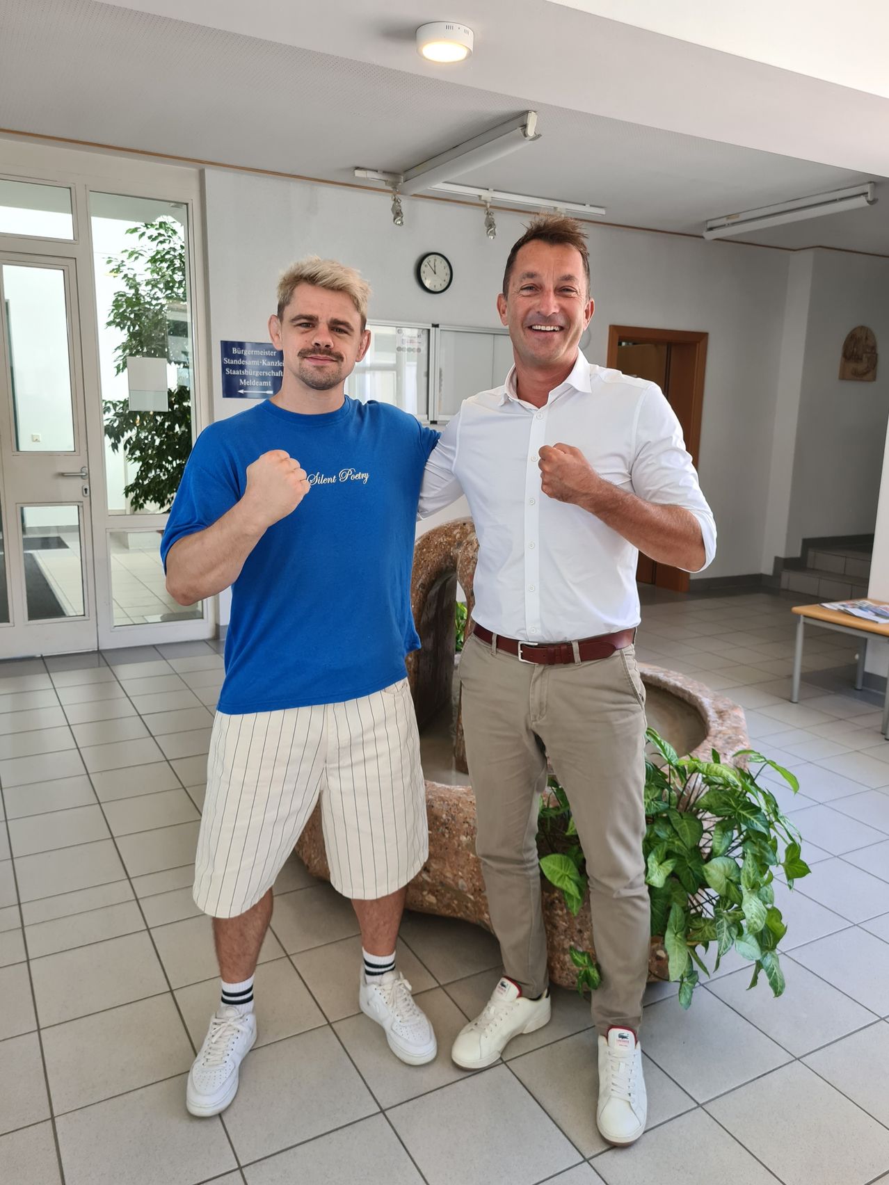 Two men are standing in a hallway, both wearing white sneakers. The man on the left is wearing a blue shirt and white shorts, while the man on the right is wearing a white shirt and khaki pants. They are both smiling and posing for a photo. Behind them is a plant and a clock on the wall.