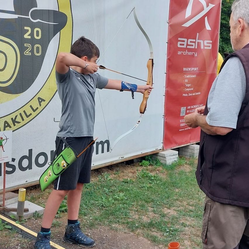 Ein junger Junge übt draußen Bogenschießen und zielt mit einem Bogen auf ein Ziel. Ein erwachsener Mann steht neben ihm und beobachtet. Im Hintergrund befindet sich ein Banner mit dem Text 'ashs archery' und einem Logo.
