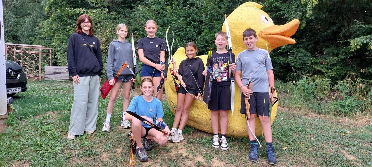 Eine Gruppe junger Bogenschützen, sowohl Jungen als auch Mädchen, posiert für ein Foto vor einer großen gelben Enten-Zielscheibe. Sie halten alle Bögen und Pfeile.
