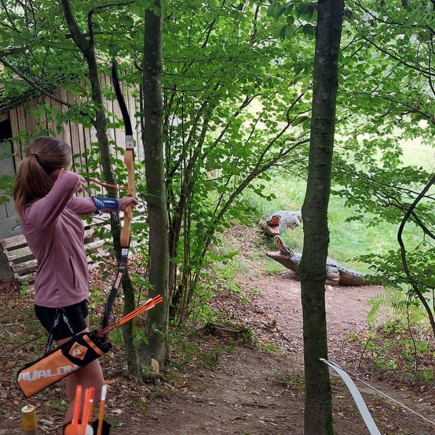 Ein junges Mädchen übt Bogenschießen im Wald, mit einer großen Alligatorstatue im Hintergrund. Sie trägt ein rosa Shirt und schwarze Shorts und hält einen Bogen und einen Pfeil.