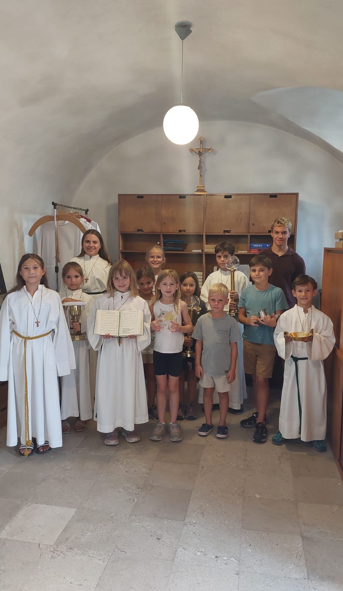 Eine Gruppe von Kindern in weißen Gewändern und mit verschiedenen Gegenständen wie einem Buch und einem Kelch stehen in einem Raum mit einem Holzregal und einem Kreuz im Hintergrund.