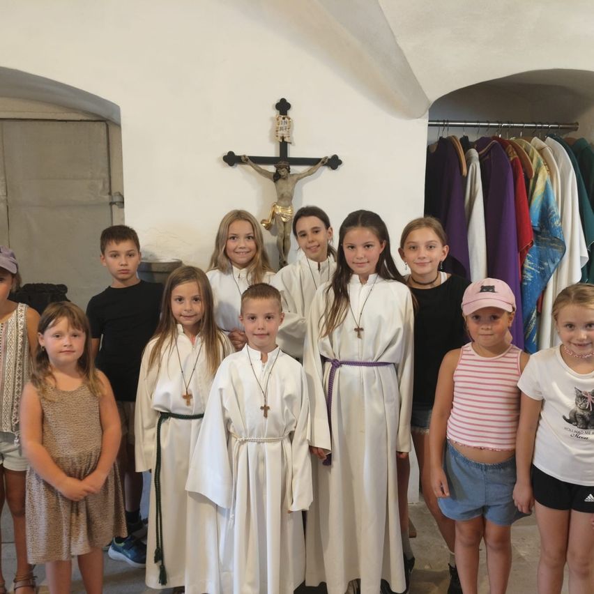 Eine Gruppe von Kindern in weißen Gewändern, einige mit Schärpen, stehen vor einem Kruzifix und einem Gestell mit farbenfrohen Gewändern in einer Kirche.