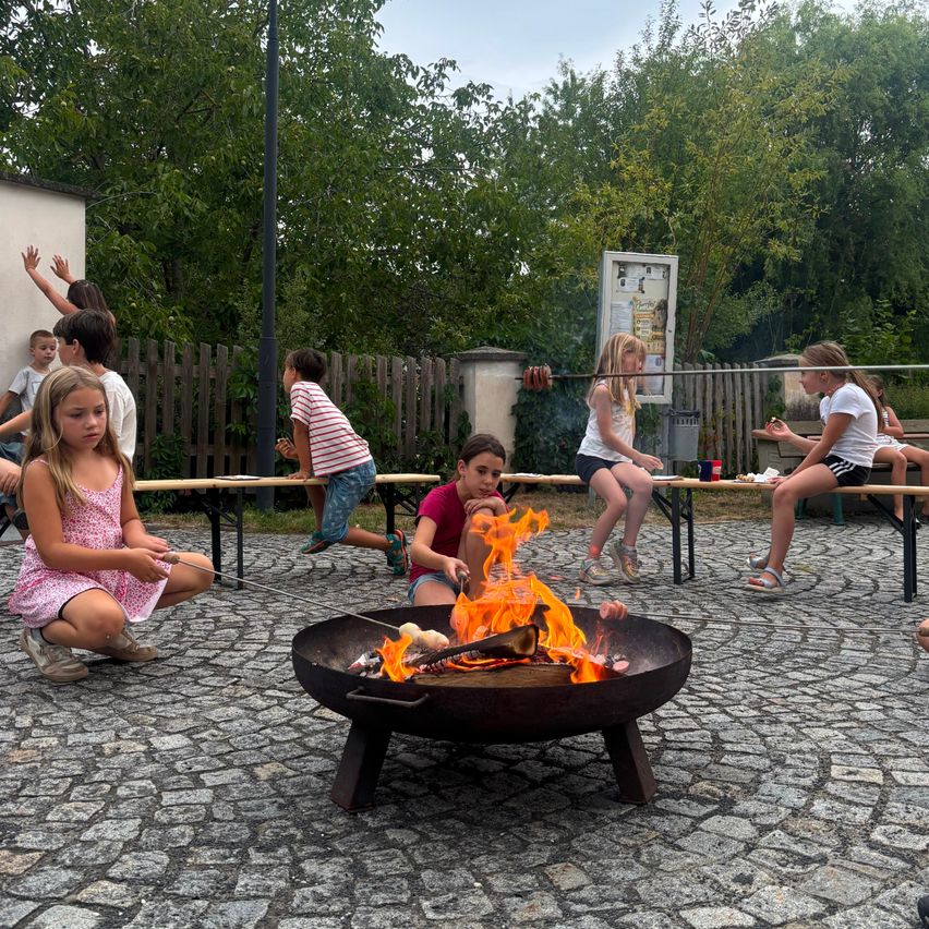 Kinder versammeln sich um ein Feuer, rösten Marshmallows auf Stöcken. Einige sitzen auf Bänken, andere stehen, mit Bäumen und einem Gebäude im Hintergrund.