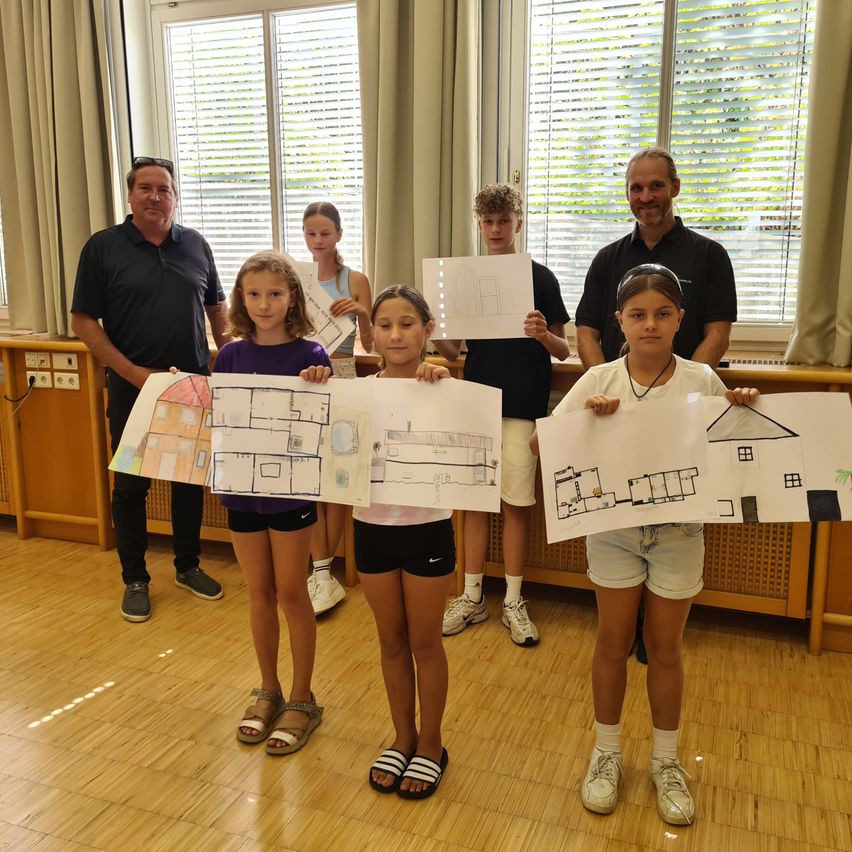 Eine Gruppe von Kindern und zwei Männern steht in einem Raum und hält Zeichnungen von Häusern. Die Kinder lächeln und scheinen für ein Foto zu posieren.