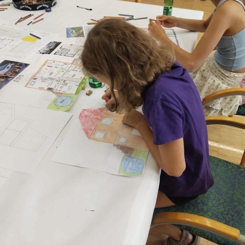 Zwei junge Mädchen sitzen an einem Tisch und malen ein Haus. Verschiedene Buntstifte und Papiere sind auf dem Tisch verstreut.