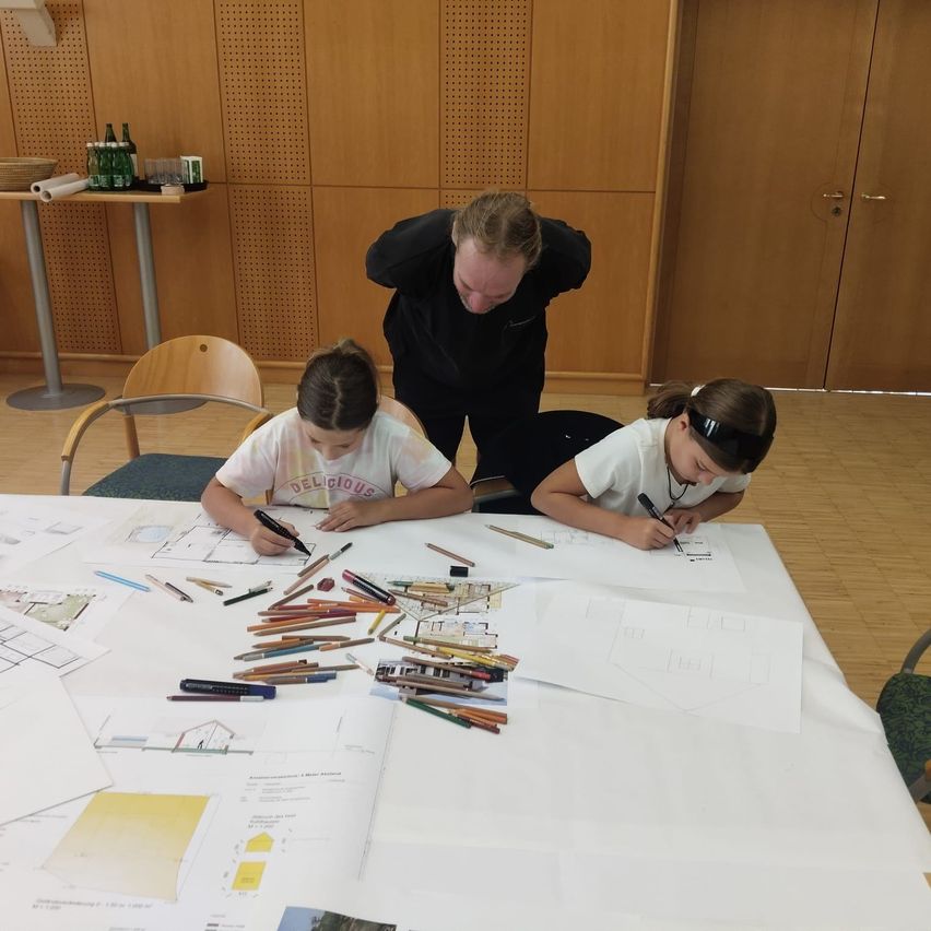 Zwei junge Mädchen zeichnen mit Buntstiften auf einem Tisch, während ein Mann hinter ihnen steht. Der Tisch ist mit Papieren und Buntstiften bedeckt.