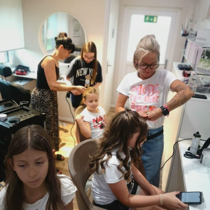 Mehrere Frauen und Mädchen in einem Salon, eine Frau frisiert die Haare eines jungen Mädchens, während ein anderes Mädchen ein Telefon in der Hand hält.