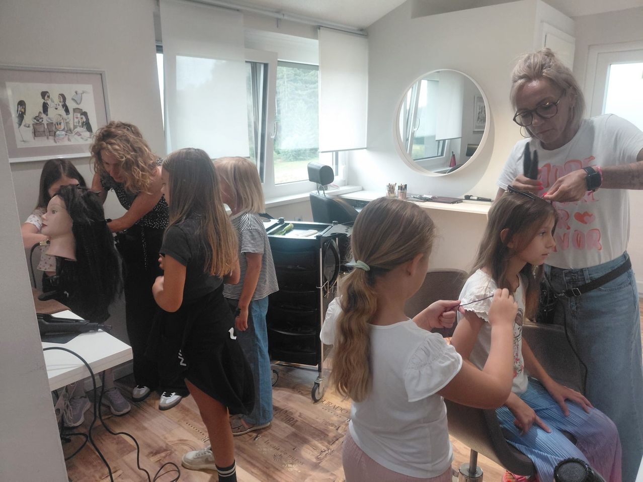 Mehrere junge Mädchen lassen sich in einem Salon die Haare stylen. Eine Frau schneidet die Haare eines jungen Mädchens. Eine andere Frau fixiert die Haare eines anderen Mädchens. Eine Frau steht und fixiert die Haare eines anderen Mädchens.