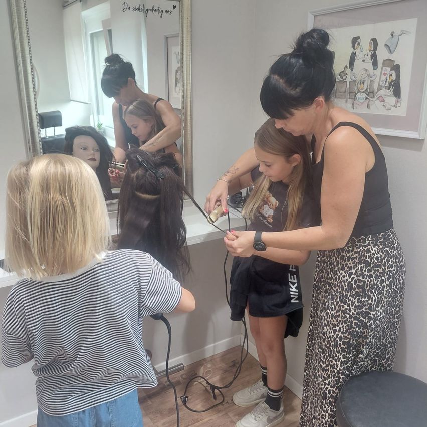 Eine Frau frisiert die Haare eines jungen Mädchens in einem Salon. Das Mädchen sitzt auf einem Stuhl, und die Frau steht hinter ihr und hält eine Haarbürste. Ein Mannequinkopf steht auf einem Tisch vor ihnen. Ein Spiegel ist an der Wand hinter ihnen montiert, und ein Bilderrahmen befindet sich rechts.