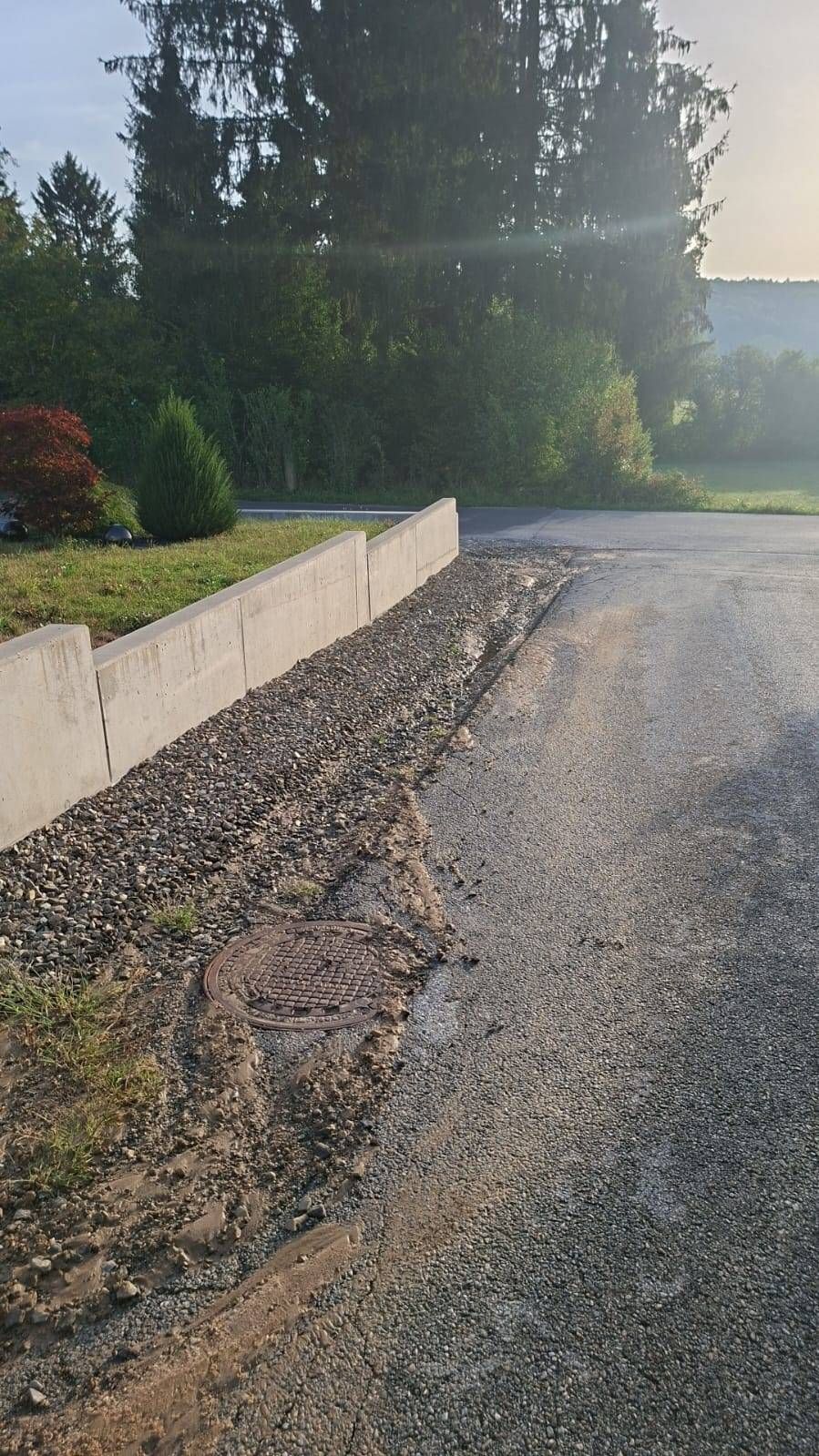 Ein Betonfundament mit Kies befindet sich neben einer Straße. Ein Schachtdeckel liegt neben der Mauer auf dem Boden.