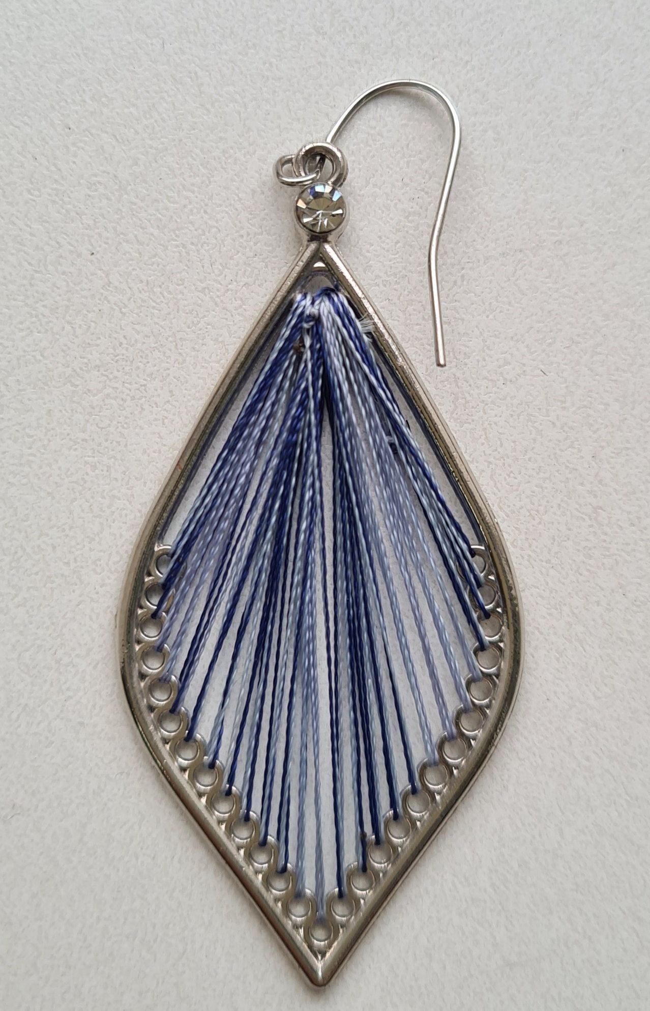 Ein silberner Ohrring mit blattförmigem Design, verziert mit blauen und weißen Fäden, die in einem geometrischen Muster gewebt sind. Der Ohrring hat einen kristallartigen oberen Teil und silberne Haken.