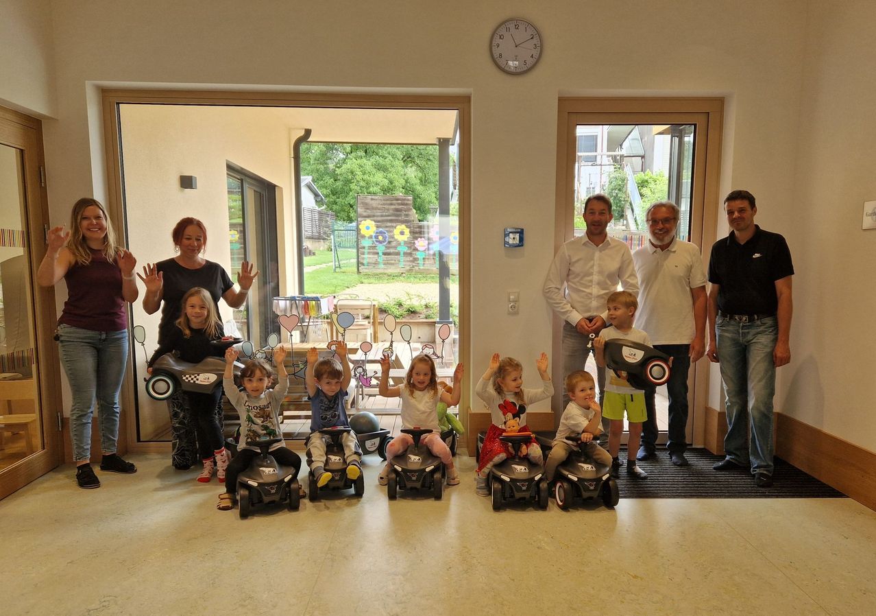 Eine Gruppe von Kindern mit kleinen Spielzeugautos posiert für ein Foto mit Erwachsenen in einem Raum mit Glastüren.