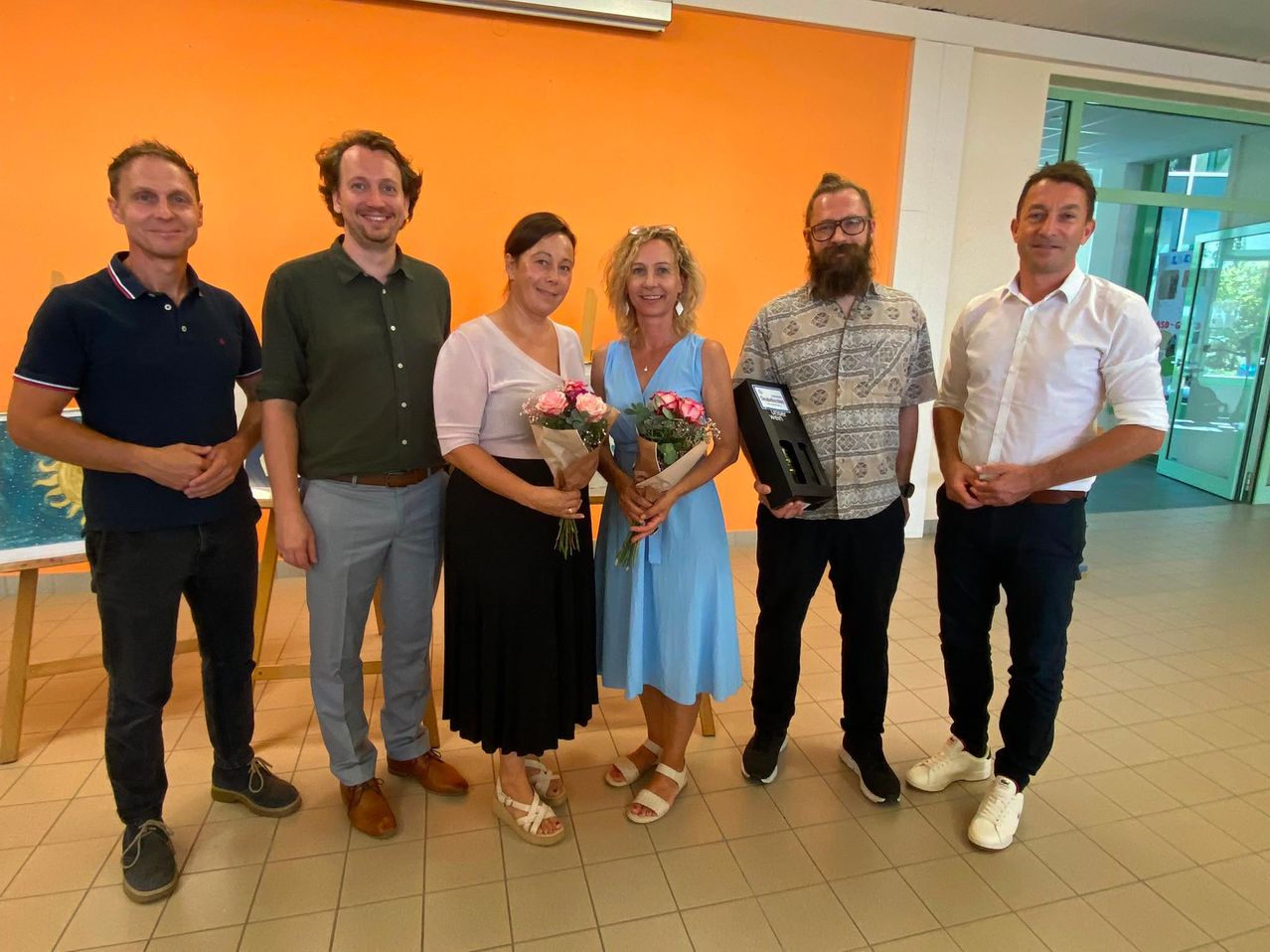 A group of six people, three women and three men, are standing together in a room. The women are holding bouquets of flowers, and one man is holding a plaque. They are all smiling and appear to be posing for a photo.