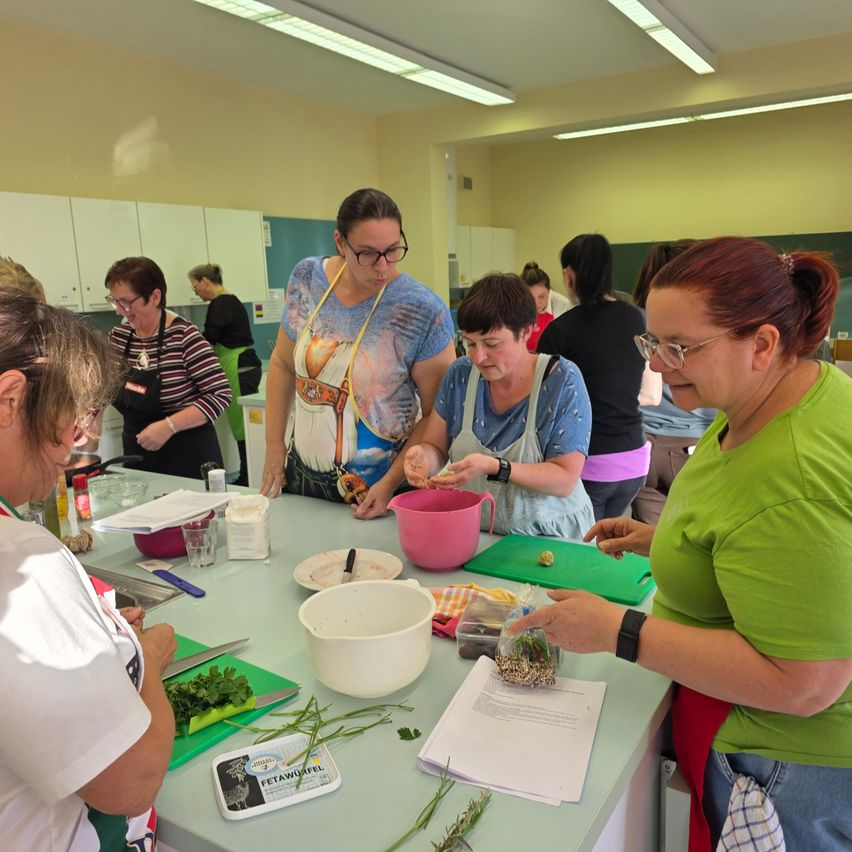 Eine Gruppe von Frauen versammelt sich in einer Küche um einen Tisch und bereitet Essen zu. Sie tragen Schürzen und haben verschiedene Zutaten und Utensilien auf dem Tisch.