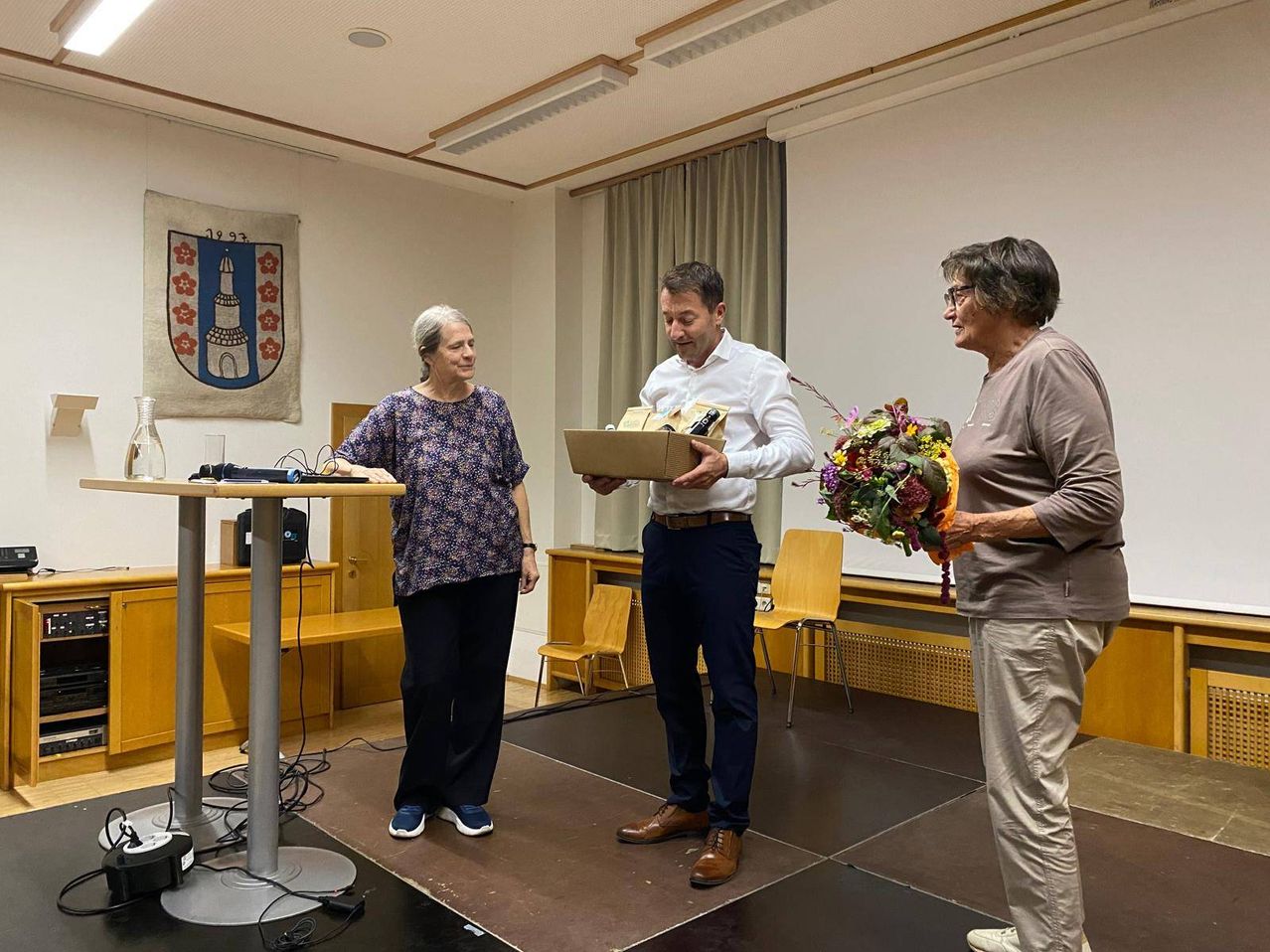 Ein Mann mit einer Schachtel und Blumen steht zwischen zwei Frauen auf der Bühne.