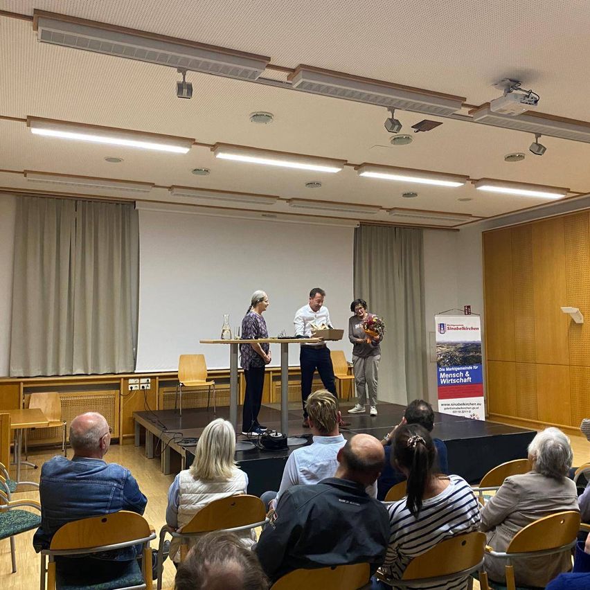 Eine Gruppe von Menschen ist in einem Raum für eine Präsentation versammelt. Drei Personen stehen auf einer Bühne mit einem Podium und einem Banner.