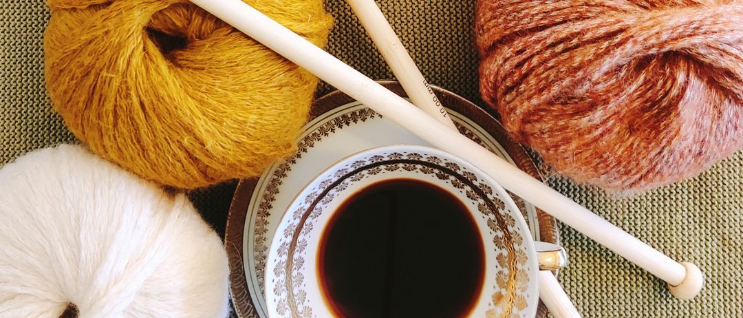 Drei Knäuel Wolle, zwei Stricknadeln und eine Tasse Kaffee auf einem Tisch. Die Wolle ist in Gelb, Orange und Weiß.