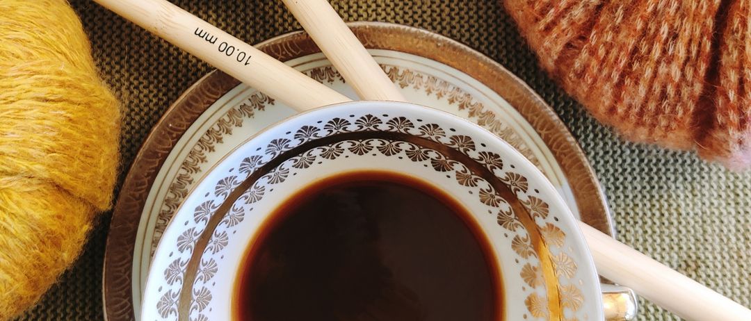 Eine Tasse Kaffee mit dekorativer Goldverzierung und zwei Holzstäbchen auf einer Untertasse mit einem passenden Teller. Ein gestrickter Kürbis und eine Wollkugel liegen ebenfalls auf dem Tisch.