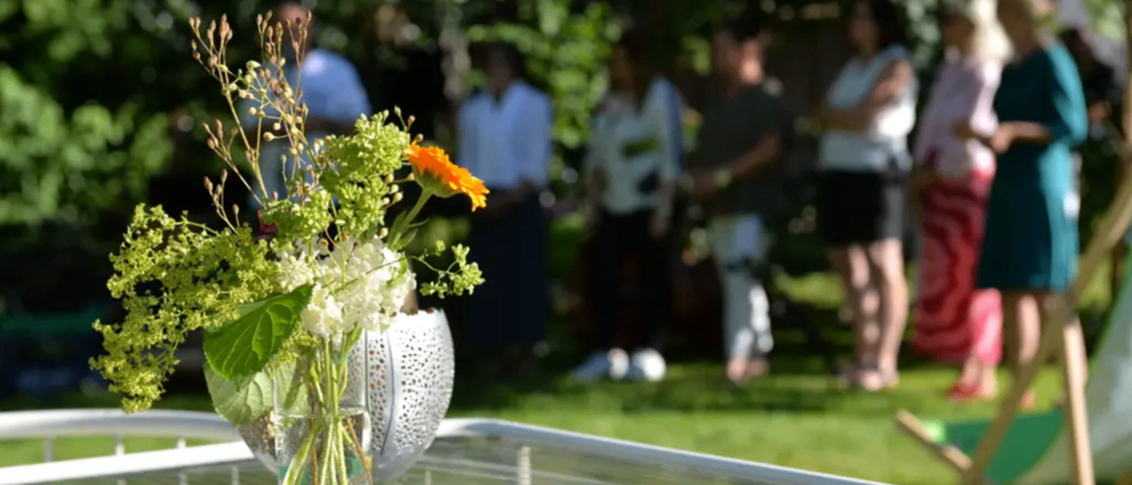 Ein Glastisch zeigt eine Vase mit Blumen. Menschen stehen in einem Garten, einige tragen Sandalen. Bäume und ein Lautsprecher sind im Hintergrund.