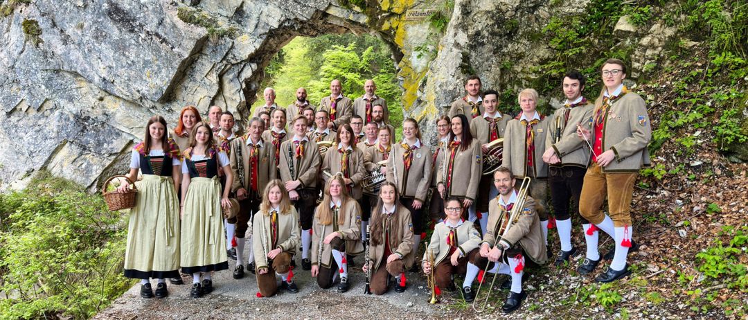 Eine Gruppe von Menschen in traditioneller Kleidung, die in einem Halbkreis vor einem felsigen Felsen stehen und sitzen. Einige halten Musikinstrumente.