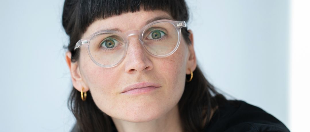 Eine Frau mit schwarzem Haar und Brille schaut geradeaus. Sie trägt ein schwarzes Shirt und hat einen Dutt. Ihre Augen sind grün, und sie posiert wahrscheinlich für ein Foto.