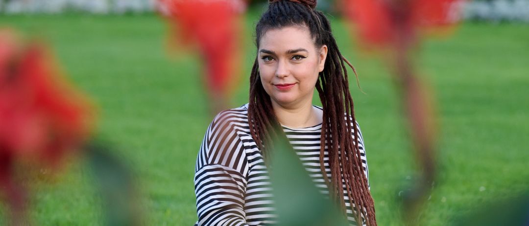 Eine Frau mit langen, geflochtenen Haaren sitzt auf einem Rasen und lächelt in die Kamera. Sie trägt ein schwarz-weiß gestreiftes Shirt. Hinter ihr bilden verschwommene rote Blumen und grüne Blätter einen farbenfrohen Hintergrund.