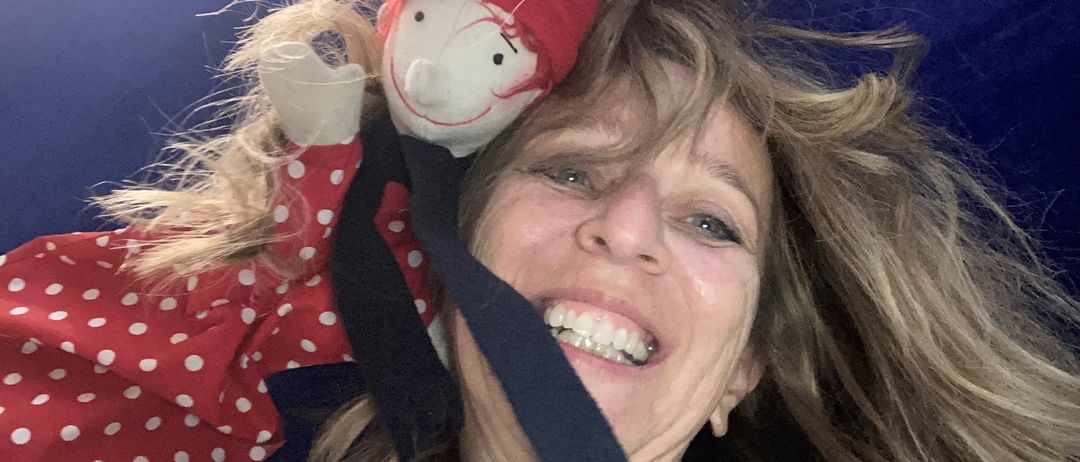 Eine lächelnde Frau mit blondem Haar hält eine Puppe auf dem Kopf, die einen roten Hut und ein rotes Punktkleid trägt. Der Hut der Puppe liegt auf ihrer Stirn.