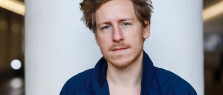 Ein Mann mit braunem Haar und Bart lächelt leicht. Er trägt eine blaue Jacke über einem weißen T-Shirt.