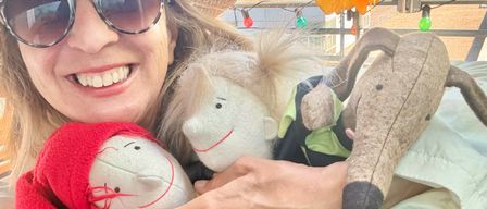 Eine Frau mit Hut und Sonnenbrille hält drei Plüschtiere, eines mit roter Kapuze, während sie auf einem Dach lächelt.