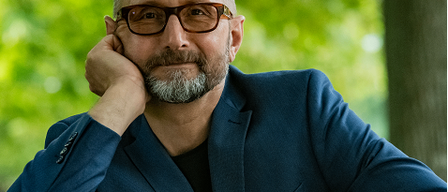 Ein Mann mit Bart und Brille, gekleidet in einen blauen Anzug, sitzt im Freien und stützt sein Kinn auf seine Hand.