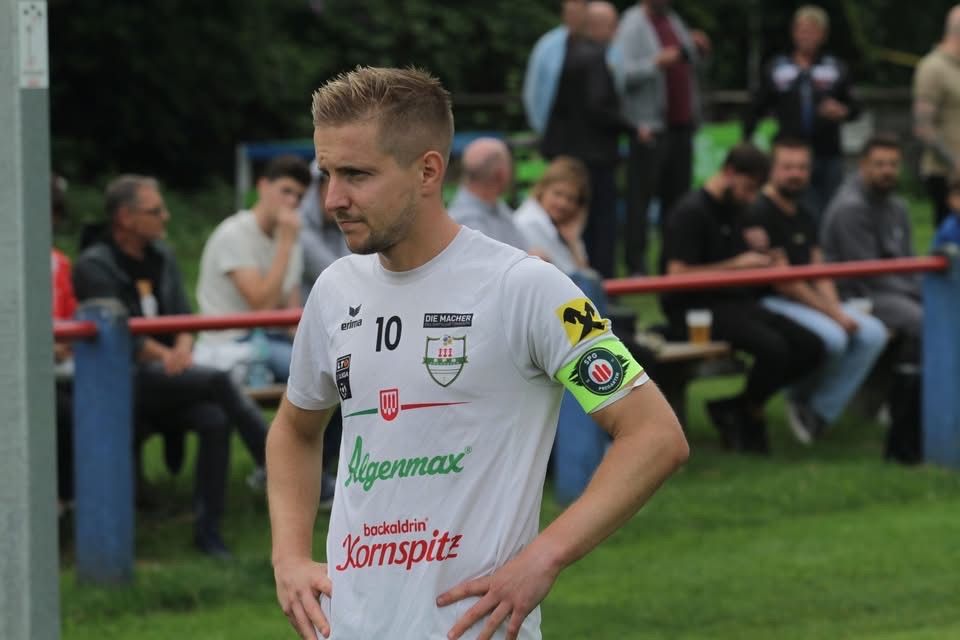 Ein Mann in einem weißen Fußballtrikot mit der Nummer 10 steht auf einem Rasenfeld, mit mehreren Personen, die auf Bänken hinter ihm sitzen.
