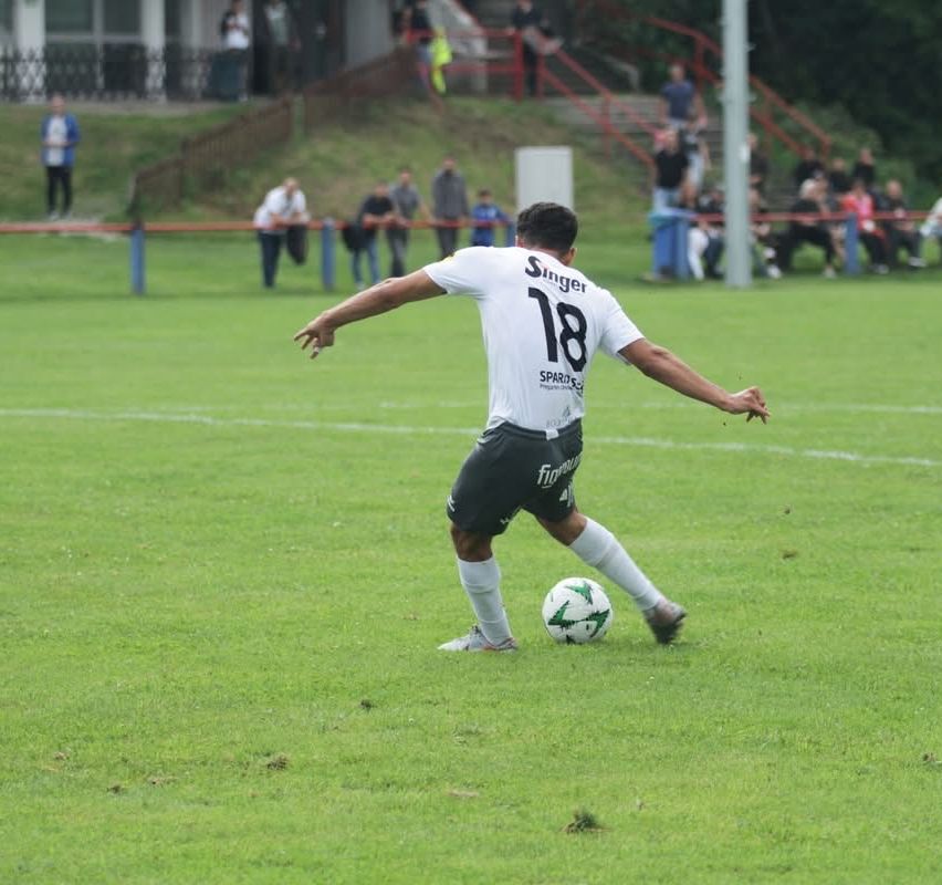 Ein Fußballspieler, der ein weißes Trikot mit der Nummer 18 trägt, tritt einen Fußball auf einem grünen Feld. Zuschauer schauen von den Tribünen zu.