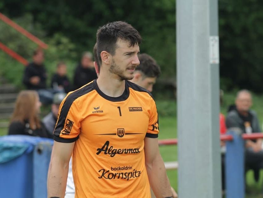 Ein Fußballspieler in einem orangefarbenen Trikot läuft über ein Feld. Er trägt ein Trikot mit der Nummer 1. Zuschauer sind im Hintergrund zu sehen.