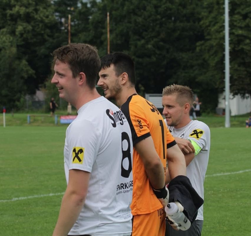 Drei Männer in Sporttrikots stehen auf einem Fußballfeld. Einer hält eine Wasserflasche. Sie scheinen zu plaudern.
