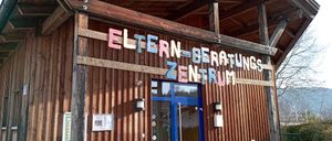 Ein holzverkleidetes Gebäude mit einem Schild, das 'Eltern-Beratungs Zentrum' besagt. Der Eingang ist blau mit Glastüren und es gibt Lampen auf beiden Seiten.
