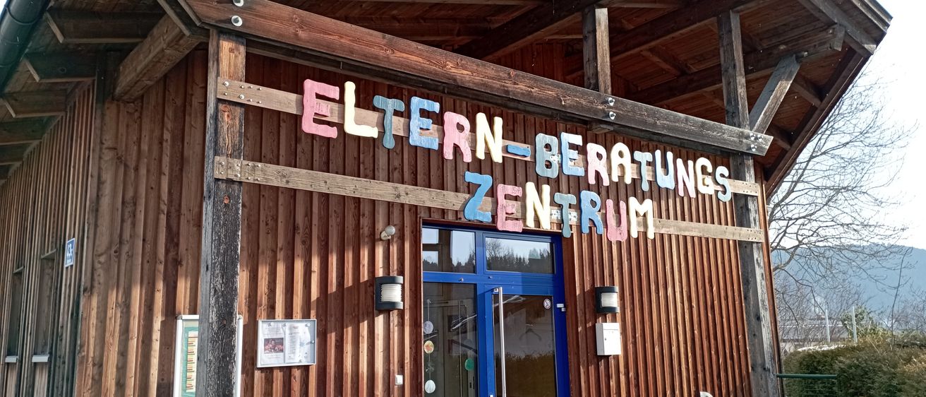 Ein holzverkleidetes Gebäude mit einem Schild, das 'Eltern-Beratungs Zentrum' besagt. Der Eingang ist blau mit Glastüren und es gibt Lampen auf beiden Seiten.