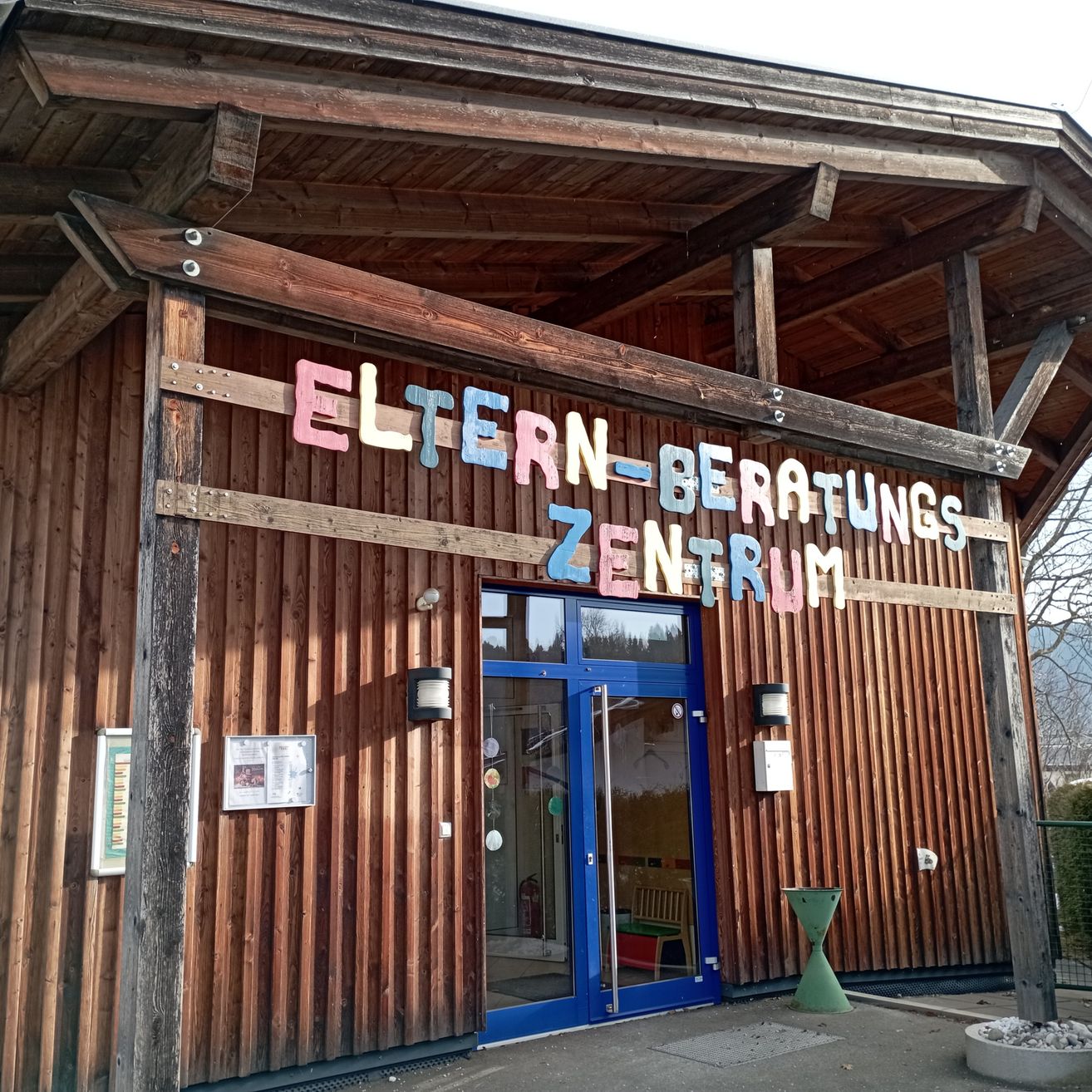 Ein hölzerne Gebäude mit einer blauen Tür, beschriftet mit 'Eltern-Beratungs Zentrum', hat dekorative Elemente, Lampen und eine Pflanze.