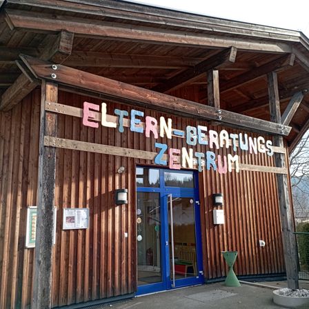 Ein hölzerne Gebäude mit einer blauen Tür, beschriftet mit 'Eltern-Beratungs Zentrum', hat dekorative Elemente, Lampen und eine Pflanze.