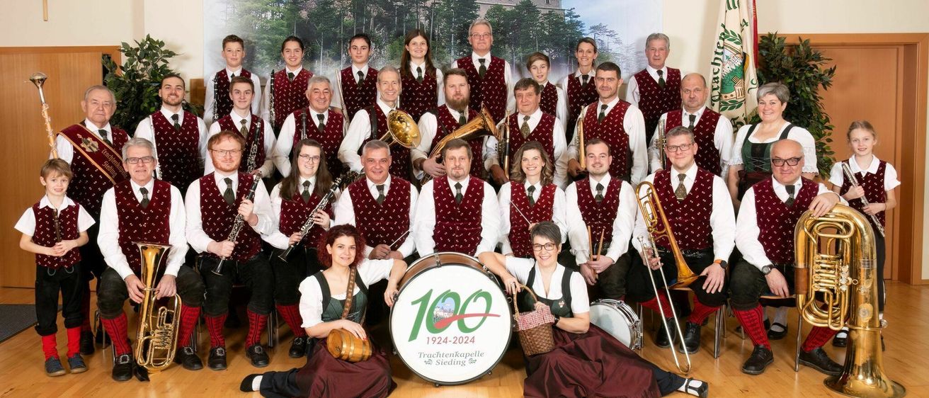 Eine Gruppe von Musikern, sowohl Männer als auch Frauen, gekleidet in traditioneller Kleidung, posieren für ein Foto mit Musikinstrumenten. Ein Banner hinter ihnen lautet '100 1924-2024 Trachtenkapelle Siedling'. Sie stehen und sitzen in einem Raum.