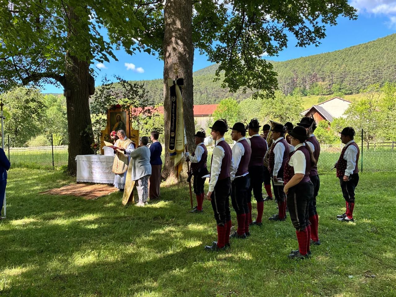 A group of people stand in a line under a tree with mountains in the background. A priest is reading from a book, and a woman holds a book.