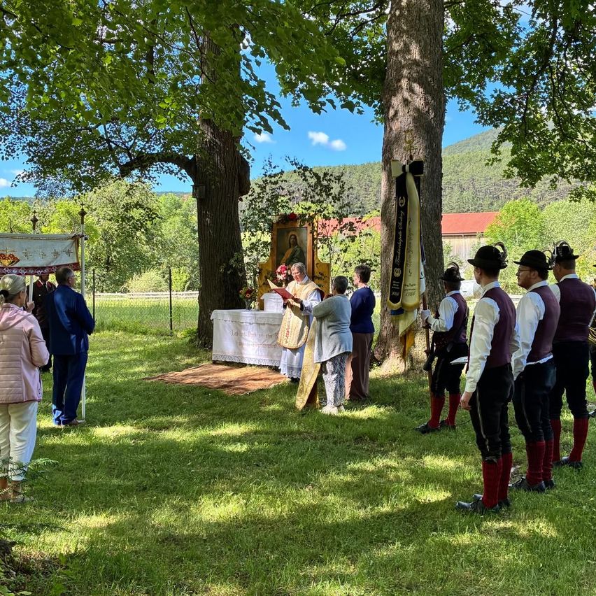 A group gathers under trees for a ceremony. A priest stands at an altar with a banner. People in traditional garb stand in formation.