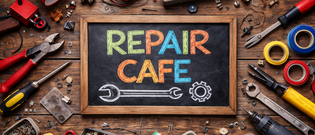 Ein Tafel mit den Worten Reparatur Café ist umgeben von verschiedenen Werkzeugen und Teilen auf einem Holztisch. Eine Lampe, ein Multimeter und ein Radio sind ebenfalls auf dem Tisch.