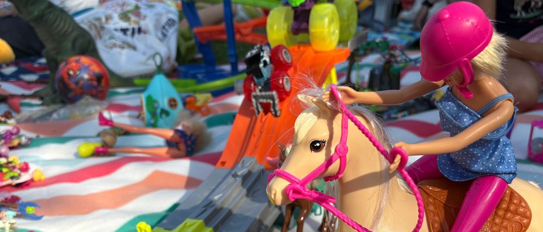 Eine Puppe reitet auf einem Pferd mit einem rosa Zügel, umgeben von mehreren Spielzeugen auf einer gestreiften Decke. Zu den Spielzeugen gehören ein Dinosaurier, ein Drache und ein Spielzeugauto.