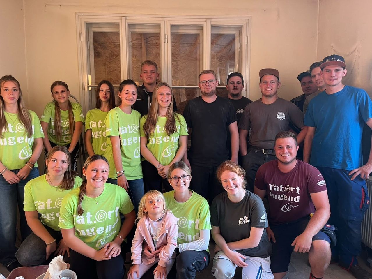 Eine Gruppe von Menschen in grünen und schwarzen T-Shirts posiert für ein Foto in einem Raum mit weißen Wänden und Fenstern.